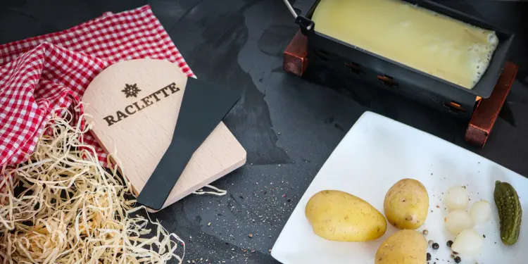 Racletteofen mit Käseschaber. Auf weissem Teller geschwellte Kartoffeln, weisse Essigzwiebeln und Cornichon
